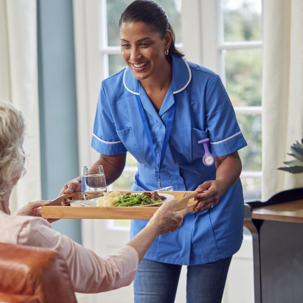 female-care-worker-in-uniform-bringing-meal-on-tray-to-senior-woman-sitting-in-lounge-at-home