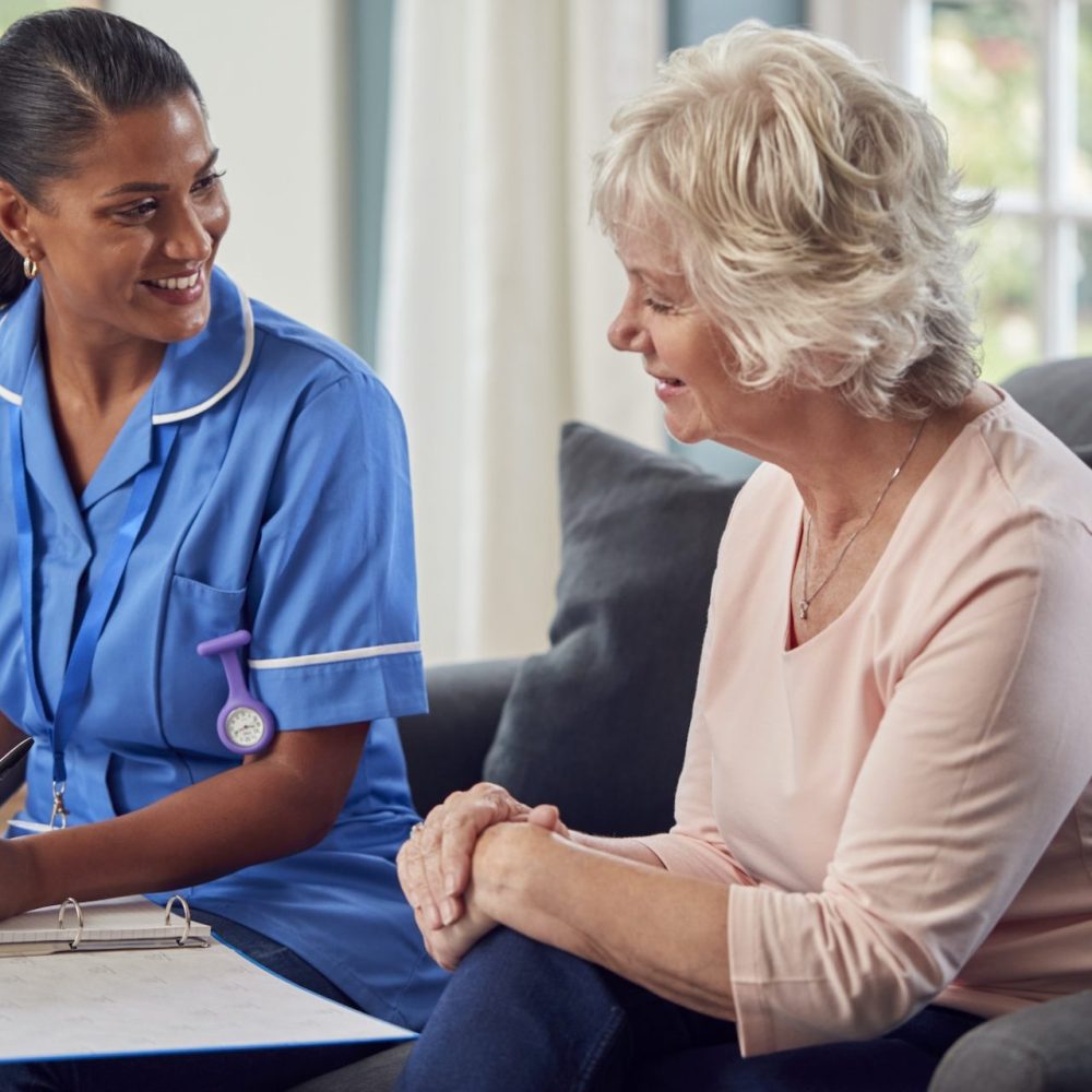 senior-woman-at-home-talking-to-female-nurse-or-care-worker-in-uniform-making-notes-in-folder-e1692669462402